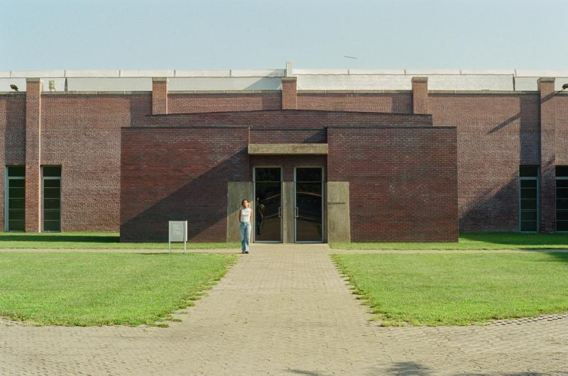 A woman poses in front of a red brick building, with a grassy front yard.