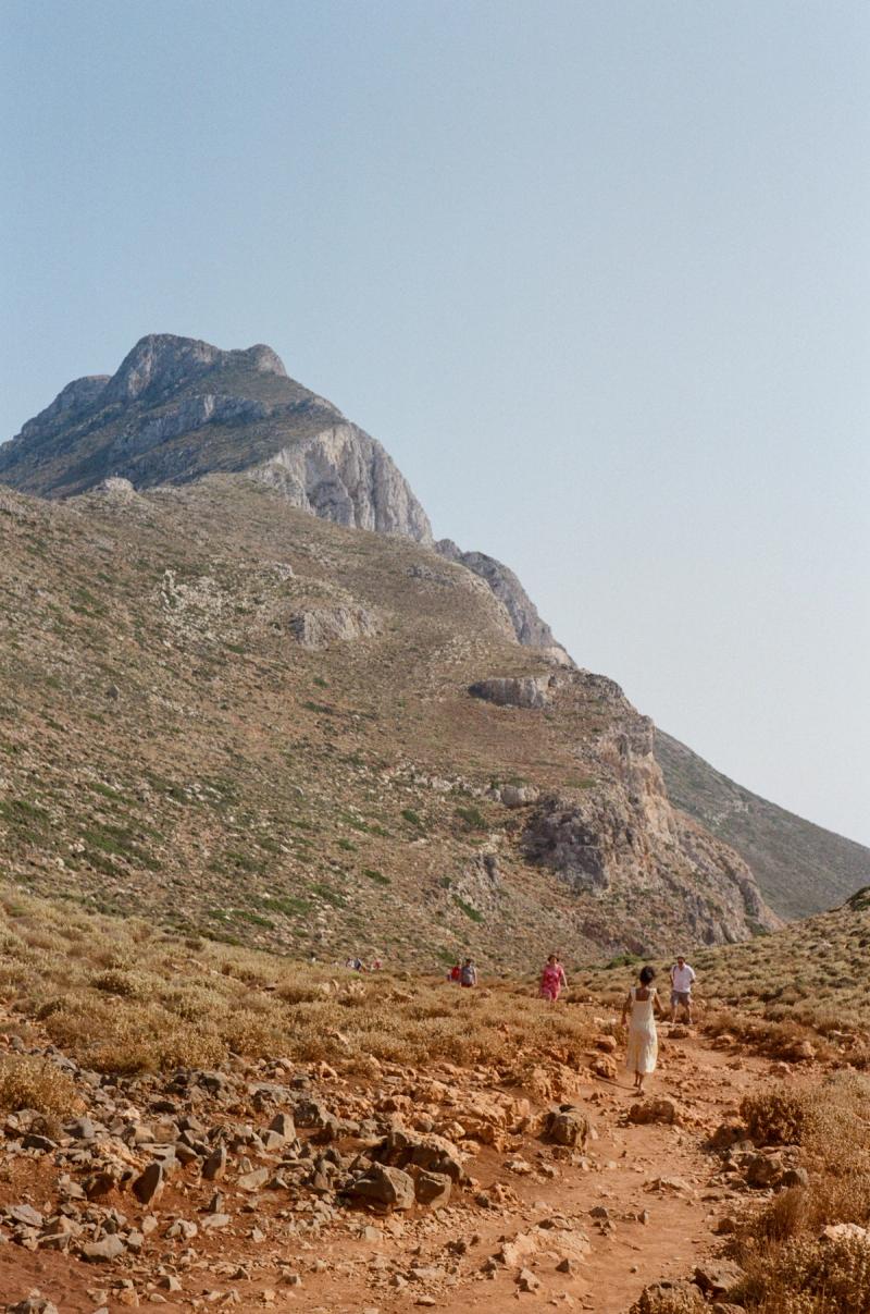 A woman in a yellow dress hikes along a dirt-covered path towards a mountain in the distance.
