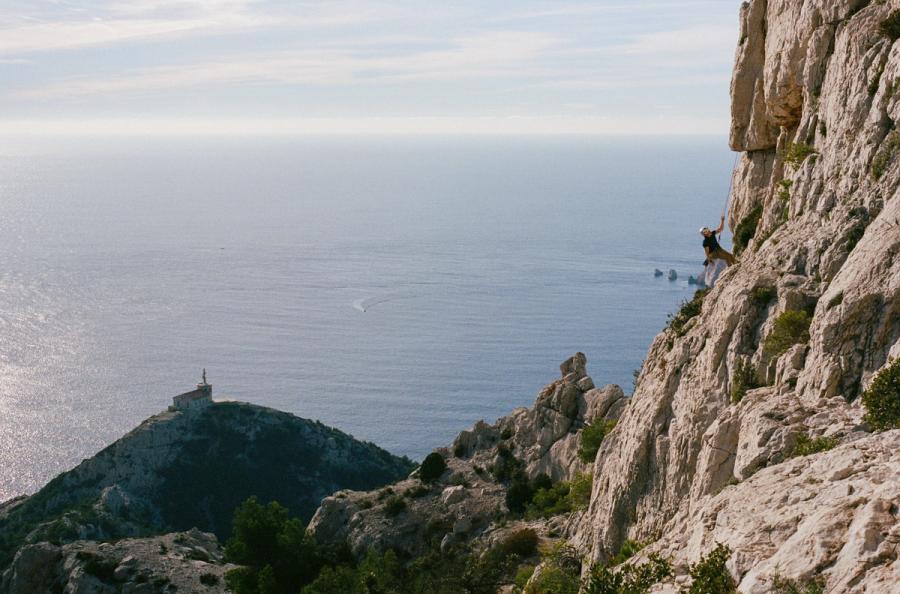 A rock climber hangs from a wall, with a church on a hill and the ocean in the background.