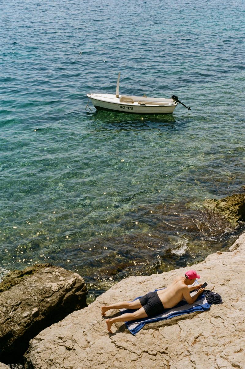 A man in a pink cap lays reading in the sun while a small boat floats in the water nearby.
