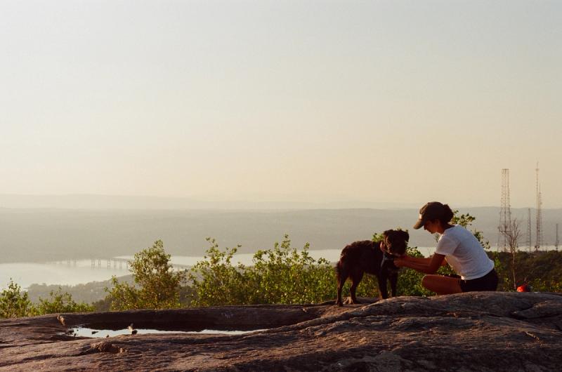 A woman adjusts her dog's harness while sitting next to a puddle.