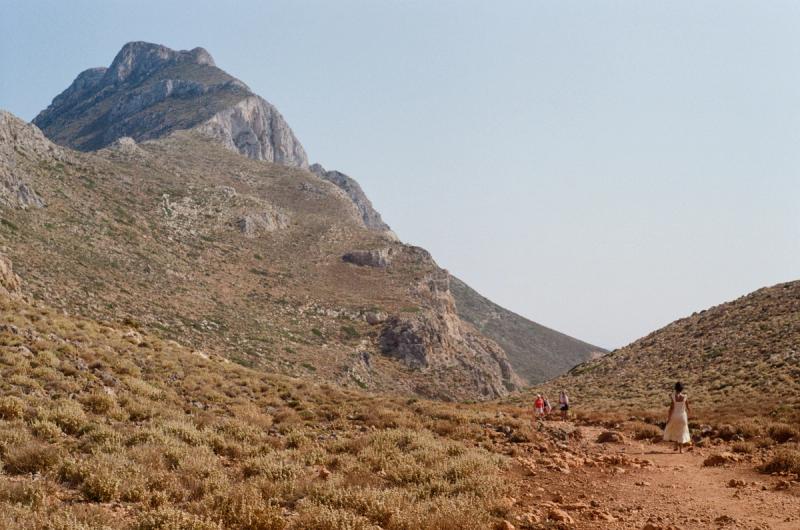 A woman in a yellow dress hikes through a dirt-covered hillside with a mountain in the distance.