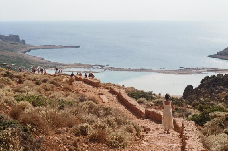 A woman in a yellow dress walks down a stone road towards a beach.