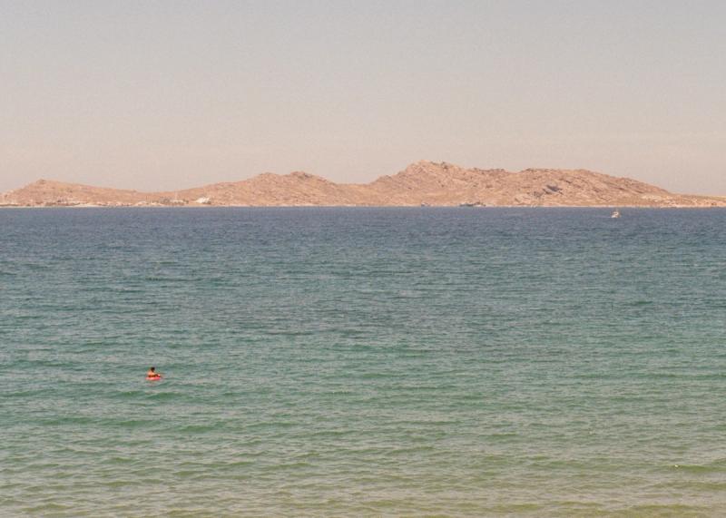 A man in a red inflatable floats in the middle of green-blue sea, staring out at the mountains in the distance.