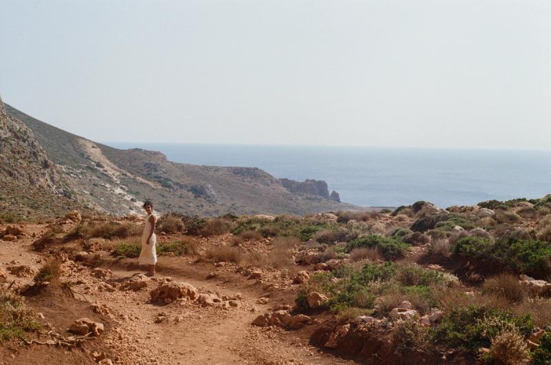 A woman in a yellow dress stares questioning into the camera, flanked by a dirt-covered hillside and a patch of blue ocean.